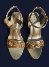 Load image into Gallery viewer, Pair of gold high-heeled sandals with decorative straps on a reflective surface.