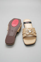 Load image into Gallery viewer, A pair of beige leather slide sandals with a decorative metallic bow embellishment on the strap. One sandal is shown from the top, displaying the beige footbed and a black brand strap with 'mmw' in white, while the other is turned to show the dark brown outsole with a red circular logo patch.