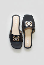 Load image into Gallery viewer, A pair of black slide sandals with metallic gold bow-shaped embellishments and rhinestone details, displayed on a textured gray stone surface.