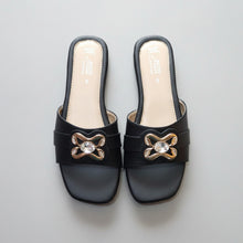 Load image into Gallery viewer, A pair of black slide sandals with a metallic gold bow-shaped ornament and a clear rhinestone in the center, displayed on a gray stone-textured surface.