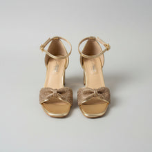 Load image into Gallery viewer, Gold high-heeled sandals with bow detail on a textured surface