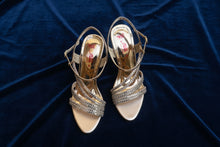 Load image into Gallery viewer, Pair of gold strappy high-heeled shoes on a gray pavement.