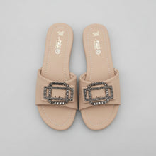 Load image into Gallery viewer, A pair of beige slide sandals with a prominent rectangular buckle embellished with black and silver rhinestones, shown on a gray textured surface.