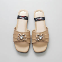 Load image into Gallery viewer, A pair of beige slide sandals with metallic silver bow embellishments and clear rhinestone centers, shown on a gray stone-textured background. The insoles feature a black band with the brand name 'milli SHOES'.