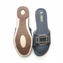 Load image into Gallery viewer, A pair of women's slide sandals in a dark slate blue color, featuring a prominent rectangular buckle embellished with rhinestones. One sandal is shown from the top view, displaying the blue footbed with gold branding, while the other is turned to show the beige and gray outsole.