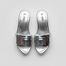 Load image into Gallery viewer, A pair of metallic silver slide sandals with a chunky square buckle detail across the strap, shown on a dark gray textured surface. The brand name 'Mill' is visible on the insole.