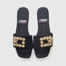 Load image into Gallery viewer, A pair of black slide sandals with a decorative gold-tone bow and floral embellishment across the strap, shown on a gray textured surface. The insole features the brand name 'milli SHOES' printed in white and red.
