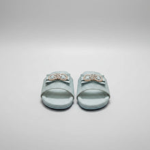 Load image into Gallery viewer, A pair of light blue glitter slide sandals with a bow and jeweled heart-shaped ornament on the strap, displayed on a gray stone surface.
