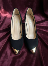 Load image into Gallery viewer, Black high-heeled shoes with gold toes on a concrete floor
