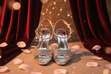 Load image into Gallery viewer, Pair of silver strappy high-heeled sandals on a reflective surface
