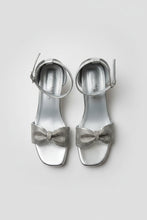 Load image into Gallery viewer, Pair of silver high-heeled sandals with bow details on a textured surface
