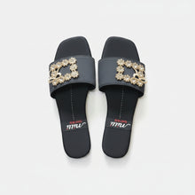 Load image into Gallery viewer, A pair of black slide sandals with decorative gold-tone floral and bow embellishments on the straps, displayed on a gray textured surface. The brand name 'milli SHOES' is visible on a black band on the footbed of each sandal.
