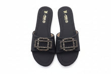 Load image into Gallery viewer, A pair of black slide sandals with large rectangular buckle embellishments covered in silver rhinestones, shown on a gray stone-textured background. The insoles display the brand name 'shuh'.
