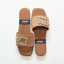 Load image into Gallery viewer, A pair of tan slide sandals with jeweled square buckle details and a small bow, shown on a gray stone-textured background. The insoles display a black brand label reading 'Milli SHOES'.
