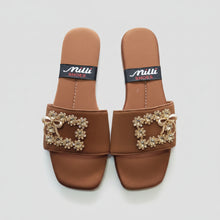 Load image into Gallery viewer, A pair of brown slide sandals with ornate gold-tone bow and floral embellishments on the straps, shown on a gray textured surface. The insoles display the brand name 'Milli SHOES'.
