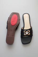 Load image into Gallery viewer, A pair of black leather slide sandals shown on a gray stone floor, with one sandal displaying the brown sole with a red heel pad and the other showing the black footbed with a metallic gold-tone bow embellishment on the strap.
