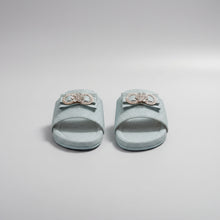 Load image into Gallery viewer, A pair of light blue glittery slide sandals with decorative metallic bow embellishments on the straps, displayed on a gray stone-textured surface.
