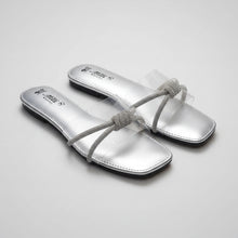 Load image into Gallery viewer, A pair of metallic silver flat slide sandals with clear plastic straps and rhinestone-embellished bow details, displayed on a dark brown textured surface. The brand name 'Milli Studio' is visible on the footbed.
