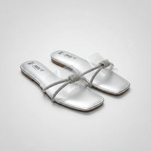 Load image into Gallery viewer, A pair of metallic silver flat slide sandals with clear straps adorned with rhinestone-knotted bows, shown on a dark textured stone surface. The brand name 'Anut B' is printed on the insoles.
