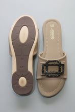 Load image into Gallery viewer, A pair of beige slide sandals with a decorative black beaded rectangular buckle on the strap, shown on a gray textured surface. One sandal is positioned to show the sole, and the other shows the insole.
